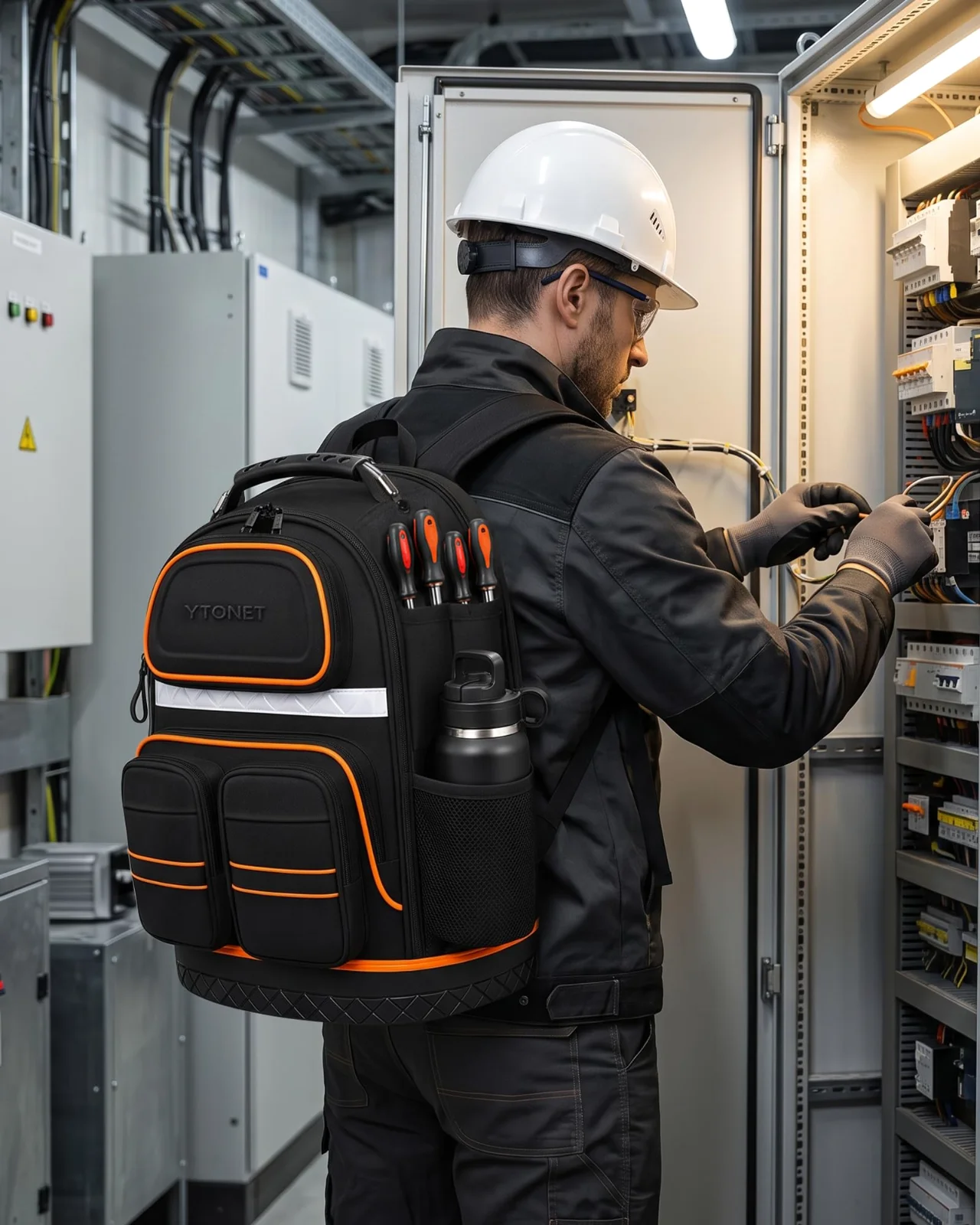 The black and orange tool backpack worn by an electrician in a hard hat at a switchboard